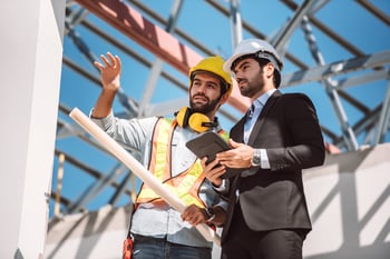 deux hommes sur un chantier. L'un d'entre eux tiens une tablette dans ses mains pendant que l'autre lui montre quelque chose au loin.