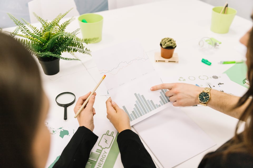 Cette image montre plusieurs personnes en réunion, examinant des graphiques représentant des données de performance. L'une des personnes utilise un crayon pour indiquer un graphique en barres tandis qu'une autre pointe un graphique linéaire. Sur la table, on trouve des objets en lien avec l'énergie renouvelable.