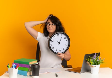 jeune-ecoliere-assez-caucasienne-anxieuse-portant-des-lunettes-est-assise-au-bureau-avec-des-outils-scolaires-met-la-main-sur-la-tete-et-tient-l-horloge-isolee-sur-l-espace-orange-avec-copie-espace