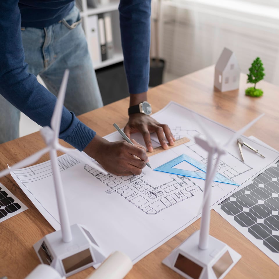 L'image représente un homme montrant un plan de construction avec son crayon. Sur la table, il y a de nombreux objets en lien avec l'environnement comme des éoliennes et des arbres.