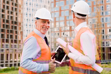 Cette image représente deux personnes avec des casque de chantier, ce qui laisse penser que ce sont des ouvriers. L'un deux tient une tablette dans sa main et fait un signe avec son pouce en l'air et il à un large sourire, ce qui laisse penser que tout s'est bien passé avec son collègue.