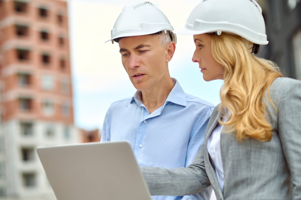 L'image montre deux professionnels du secteur du BTP, un homme et une femme, portant des casques de sécurité, l'un d'eux tenant un ordinateur portable. En arrière-plan, il y a un bâtiment en construction qui symbolise le chantier. Les deux personnes ont l'air concentré et en discussion professionnelle.