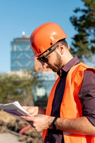 Un homme en train de regarder un papier sur un chantier. Il est habillé en ouvrier avec un casque de chantier sur la tête et une veste orange de chantier.